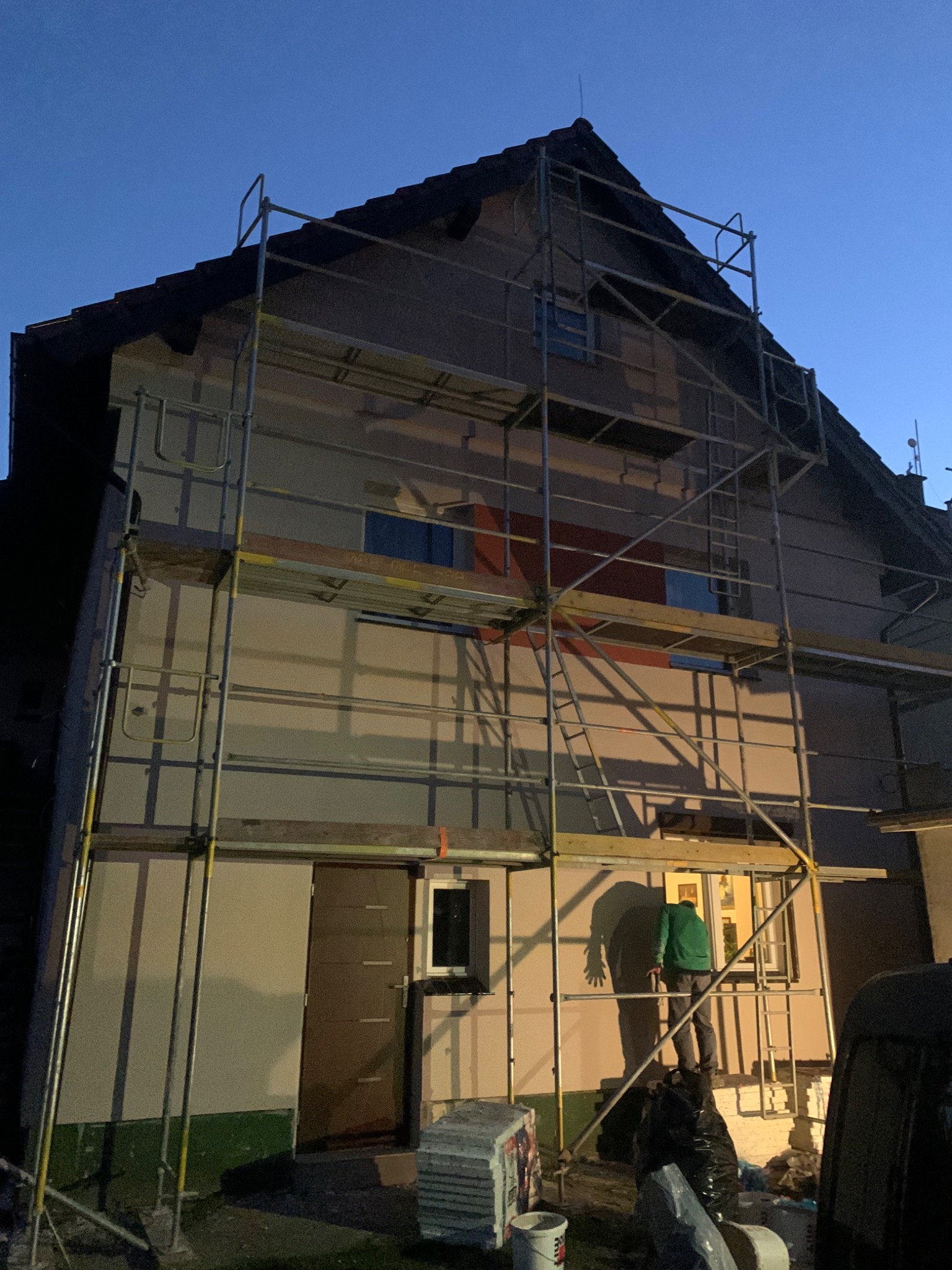 Dom w rusztowaniach podczas prac elewacyjnych o zmierzchu. Widoczny pracownik w zielonej bluzie na rusztowaniu. Częściowo otynkowana ściana.