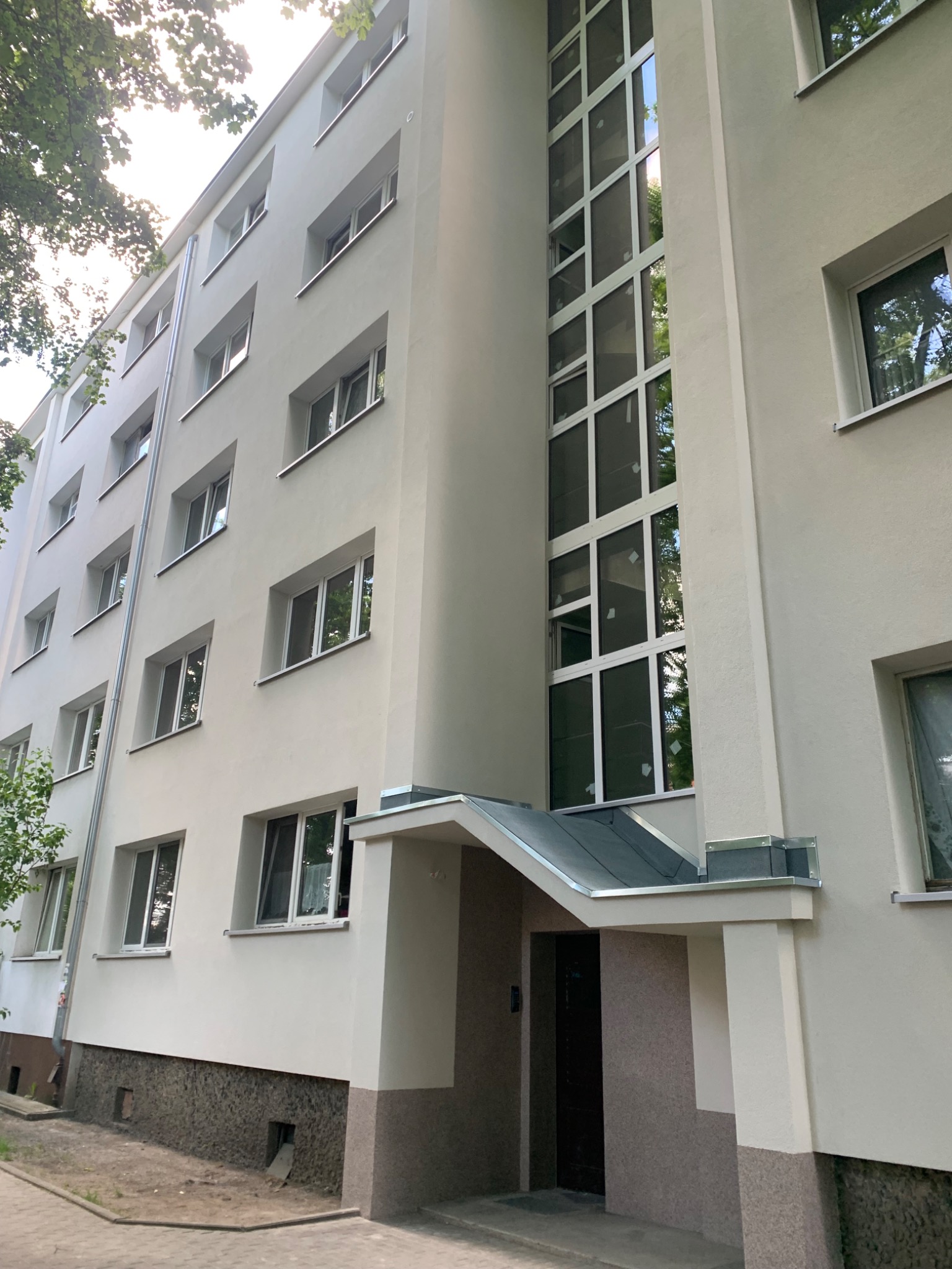 Odnowiona elewacja bloku mieszkalnego z jasną fasadą, białymi oknami i przeszkloną klatką schodową, sfotografowana z dołu w słoneczny dzień.