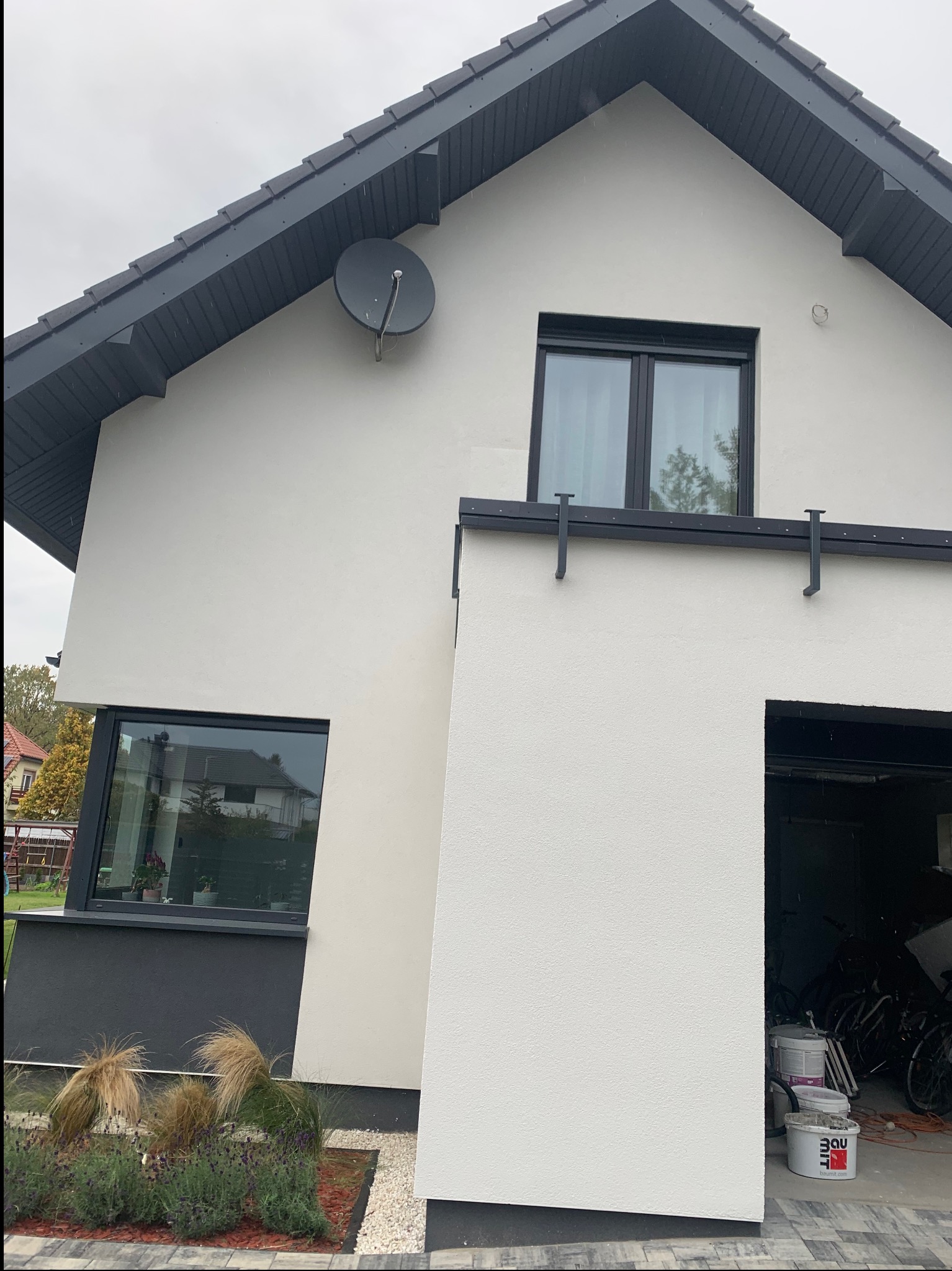 Biała elewacja domu jednorodzinnego z czarnymi ramami okiennymi i dachem. Widoczna antena satelitarna i fragment garażu z farbami. Nowoczesny styl.