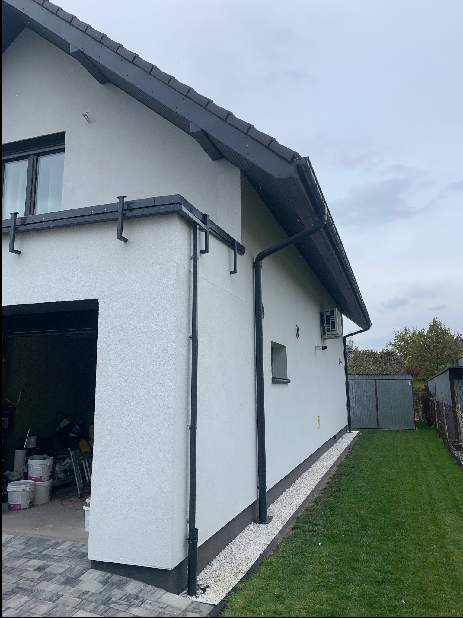 Jasna elewacja domu jednorodzinnego z czarnymi rynnami i dachem, widok z boku na tle zielonego trawnika i pochmurnego nieba. W tle widoczny garaż z akcesoriami.