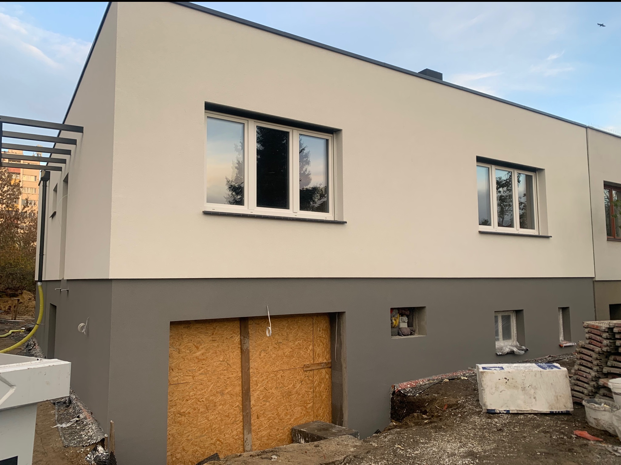 Dwukolorowa elewacja domu z widocznymi oknami i elementami konstrukcyjnymi. Dolna część w szarym kolorze, górna w jasnym odcieniu beżu. Widoczne elementy rusztowania.