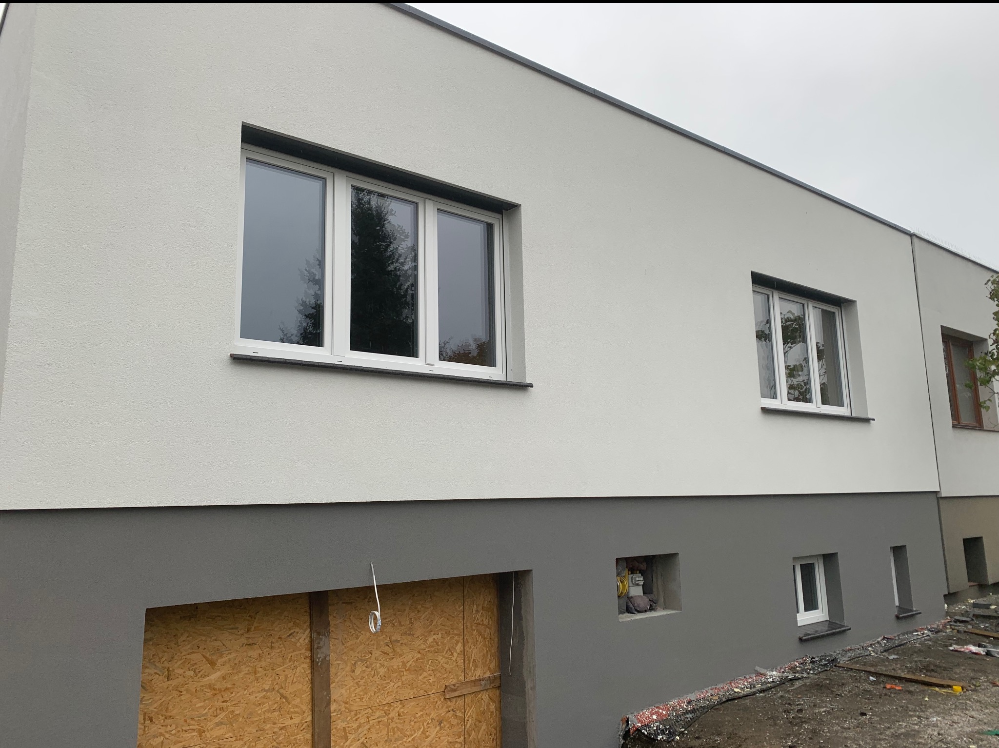 Dwukolorowa elewacja domu z białym tynkiem na górze i szarym cokołem. Okna z białymi ramami. Część cokołu zakryta płytą OSB. Widoczne ślady prac wykończeniowych.
