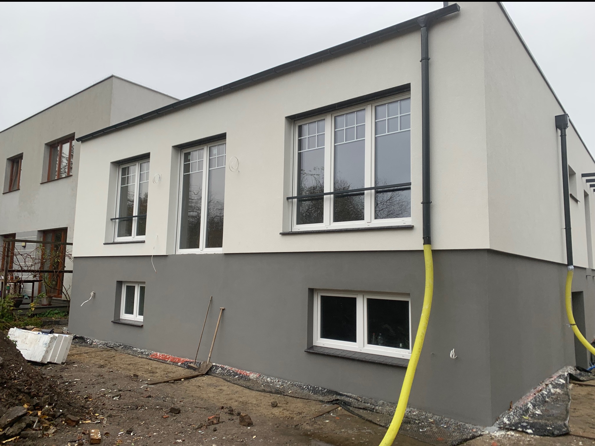 Dwukondygnacyjny dom po tynkowaniu elewacji, z widocznymi oknami z białymi ramami i szarą dolną częścią elewacji. Na ziemi leżą materiały budowlane i narzędzia.