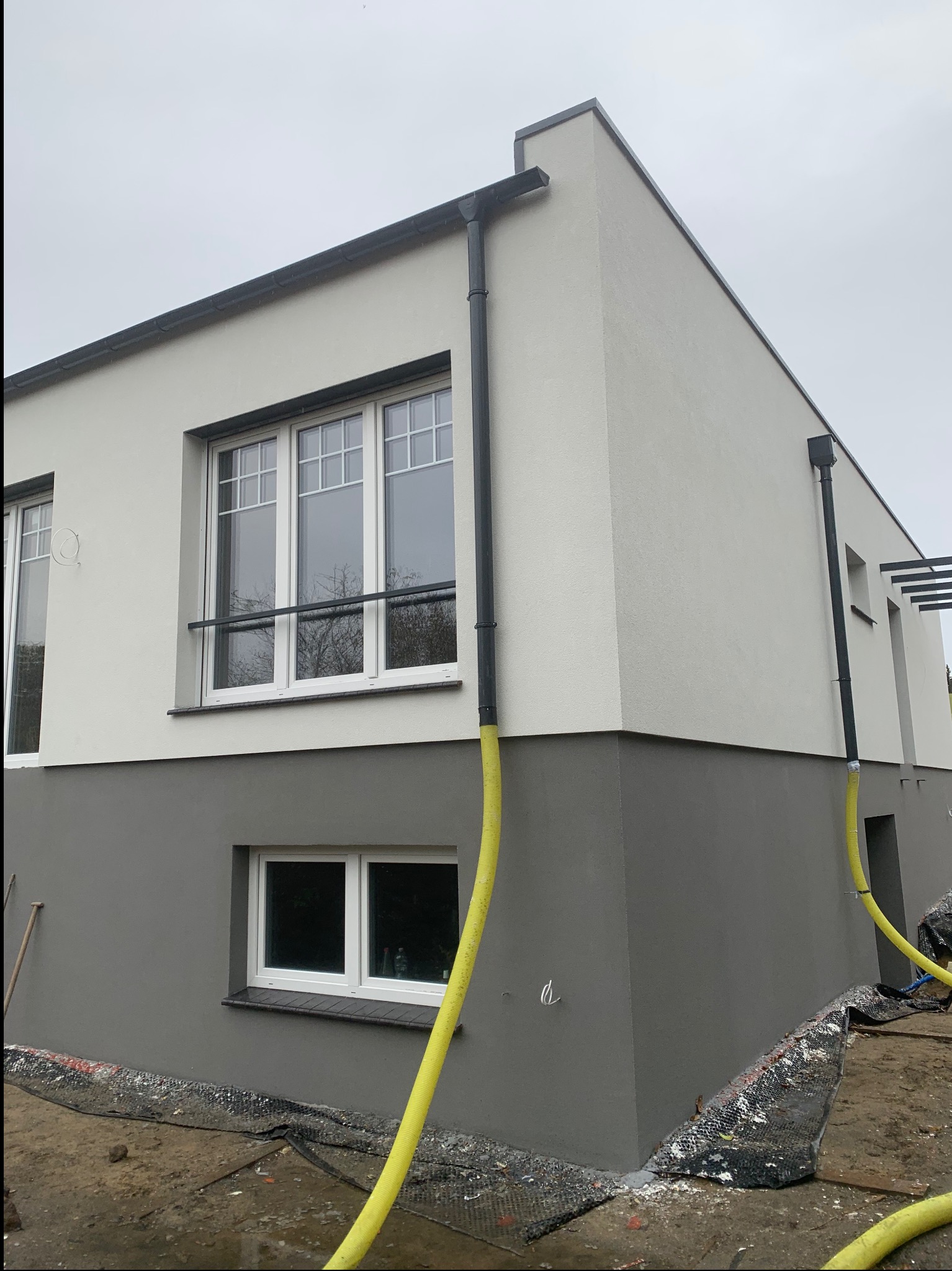 Dwukolorowa elewacja domu z widocznymi rynnami i żółtymi wężami na zewnątrz. Dolna część elewacji szara, górna biała. Widoczne okna z białymi ramami.