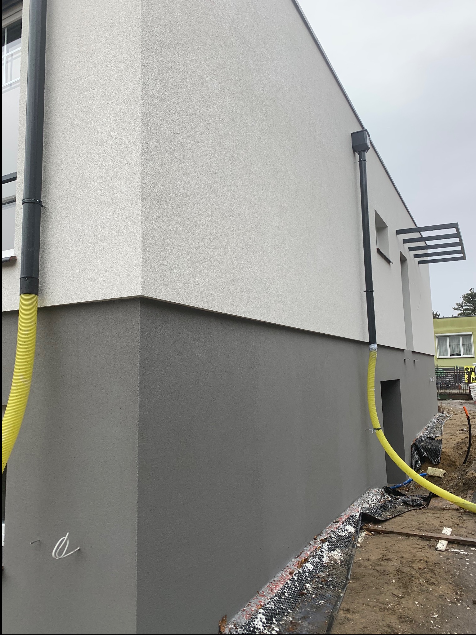 Dwukolorowa elewacja budynku z widoczną rurą spustową i żółtym wężem. Szara dolna część, biała górna. Widoczne elementy rusztowania i teren wokół budynku.