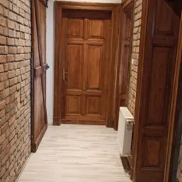 Korytarz po renowacji: ceglana ściana, drewniane drzwi przesuwne, ościeżnice z odnowioną emalią, podwieszany sufit i panele podłogowe w jasnym odcieniu.