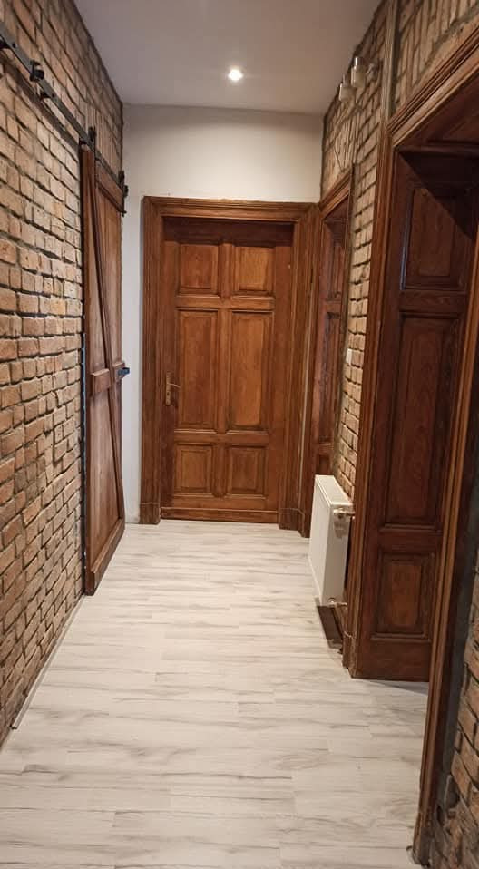 Korytarz po renowacji: ceglana ściana, drewniane drzwi przesuwne, ościeżnice z odnowioną emalią, podwieszany sufit i panele podłogowe w jasnym odcieniu.