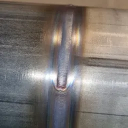 Spoina na metalowej rurze, widoczne tęczowe zabarwienie termiczne. Precyzyjne łączenie elementów, wysoka jakość wykonania. Detal spawalniczy.