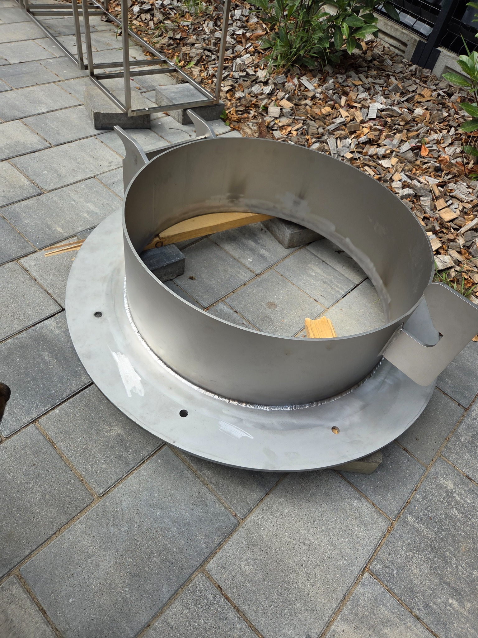 Metalowy element konstrukcyjny z widocznymi spawami, umieszczony na kostce brukowej, z drewnianymi elementami wewnątrz i metalową konstrukcją w tle.