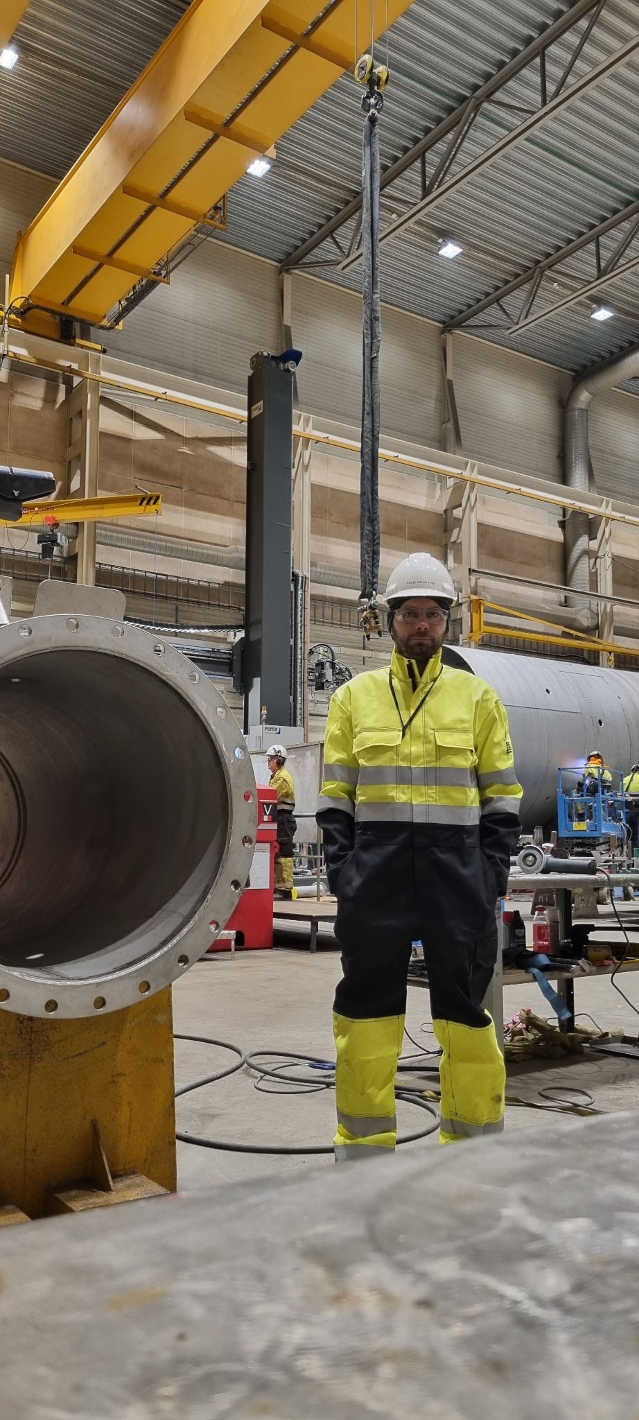 Pracownik w kombinezonie BHP stoi na hali obok spawanej rury z kołnierzem, pod suwnicą. W tle widoczni inni pracownicy w trakcie pracy.