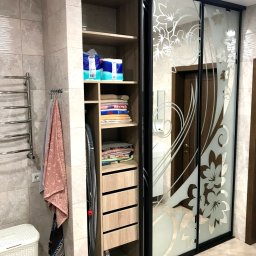 DENYS ZHABITENKO - Szafa wnękowa z przesuwnymi drzwiami zdobionymi motywem kwiatowym, w łazience z ręcznikami i grzejnikiem drabinkowym, widok na wnętrze szafy z półkami i szufladami.