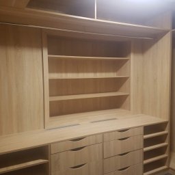 DENYS ZHABITENKO - Jasna, drewniana szafa wnękowa z szufladami i półkami, oświetlona od góry. Widoczna faktura drewna i metalowe detale wentylacyjne na blacie.