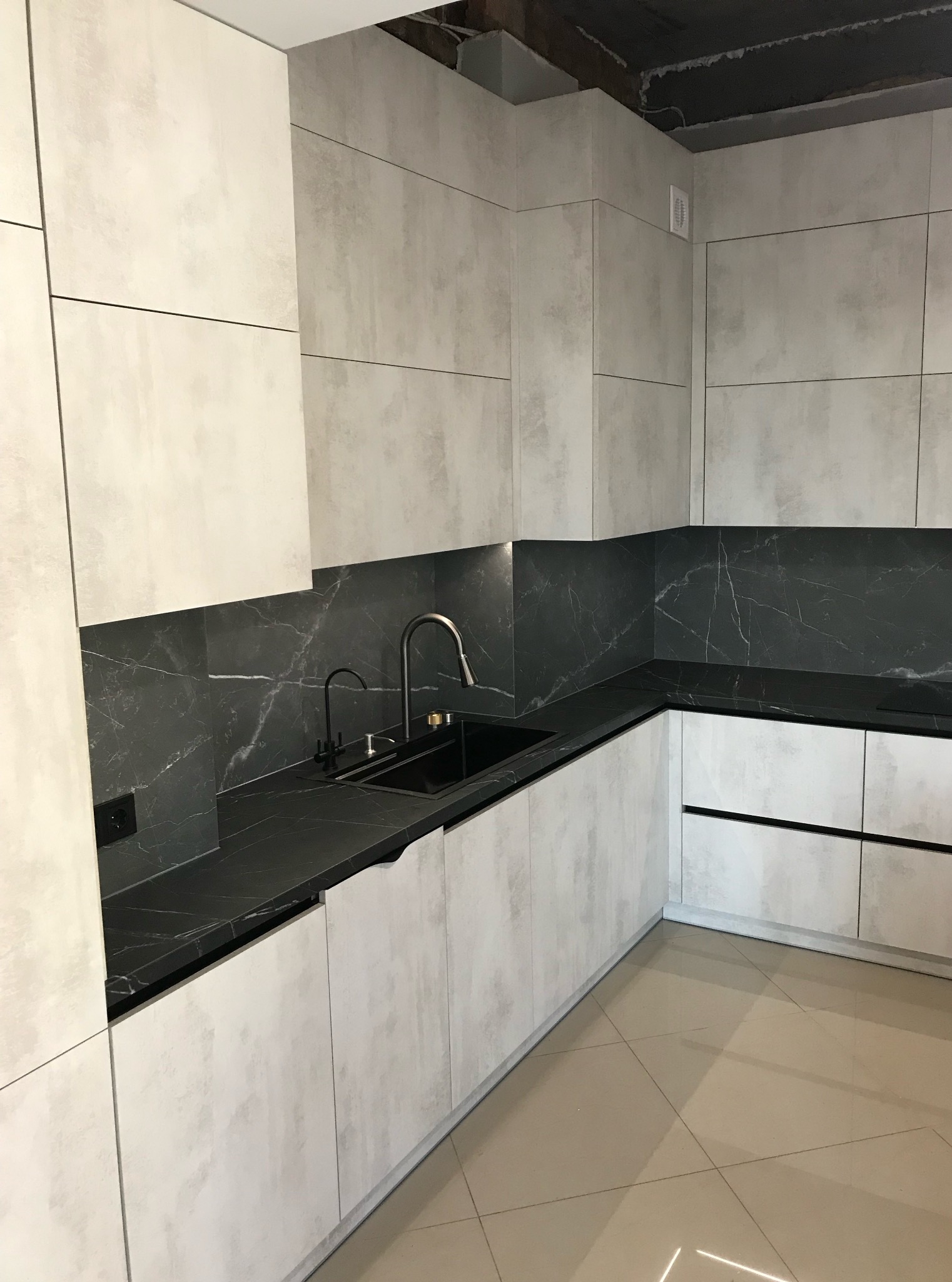 Nowoczesna kuchnia z szarymi frontami i czarnym blatem z motywem marmuru. Dwie czarne baterie przy zlewie. Widoczne płytki podłogowe w beżowym odcieniu.