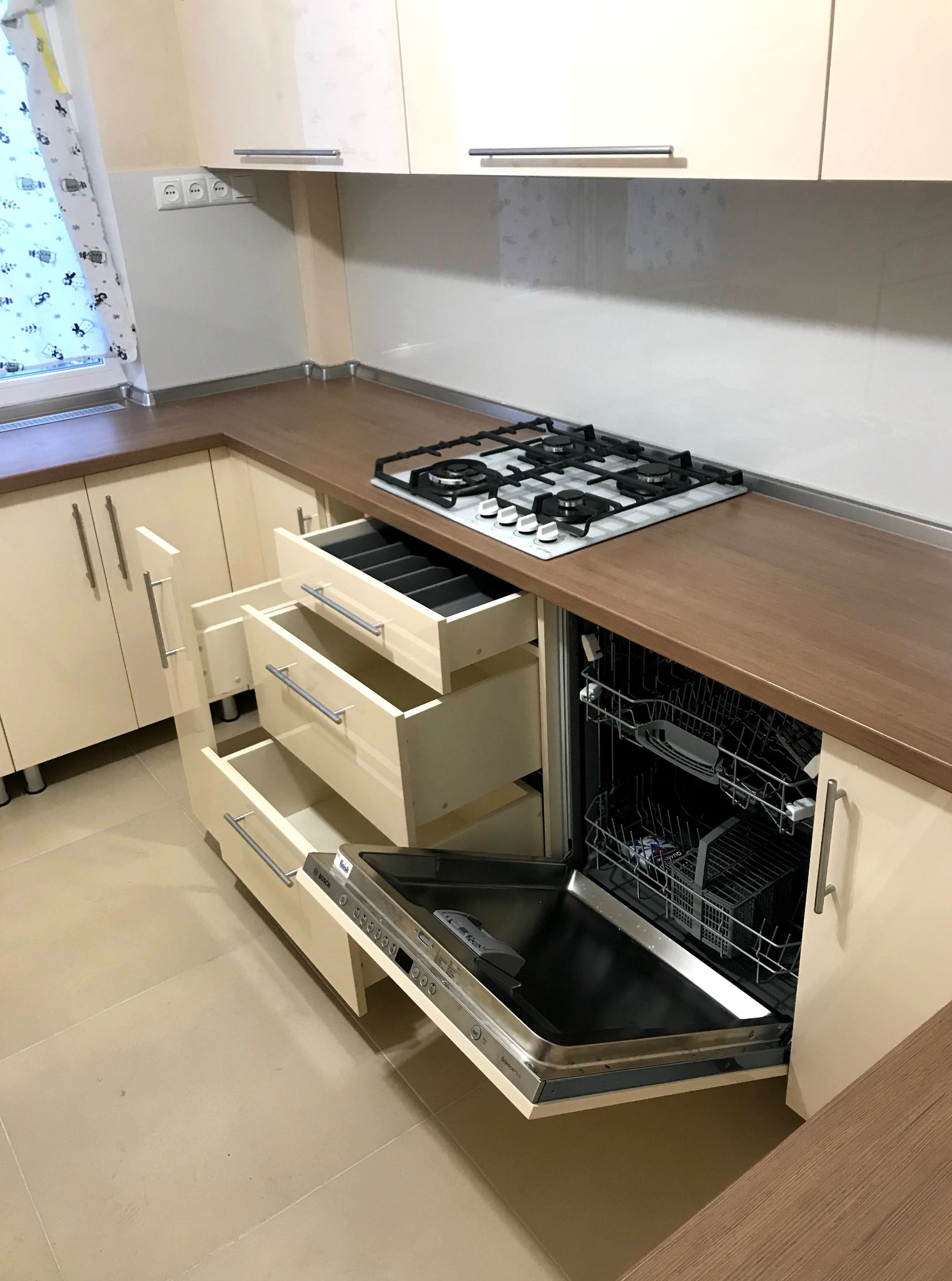 Jasna kuchnia z drewnianym blatem, otwartymi szufladami z przegródkami na sztućce i otwartą zmywarką do naczyń. Widoczna płyta gazowa i okno z zasłonką.