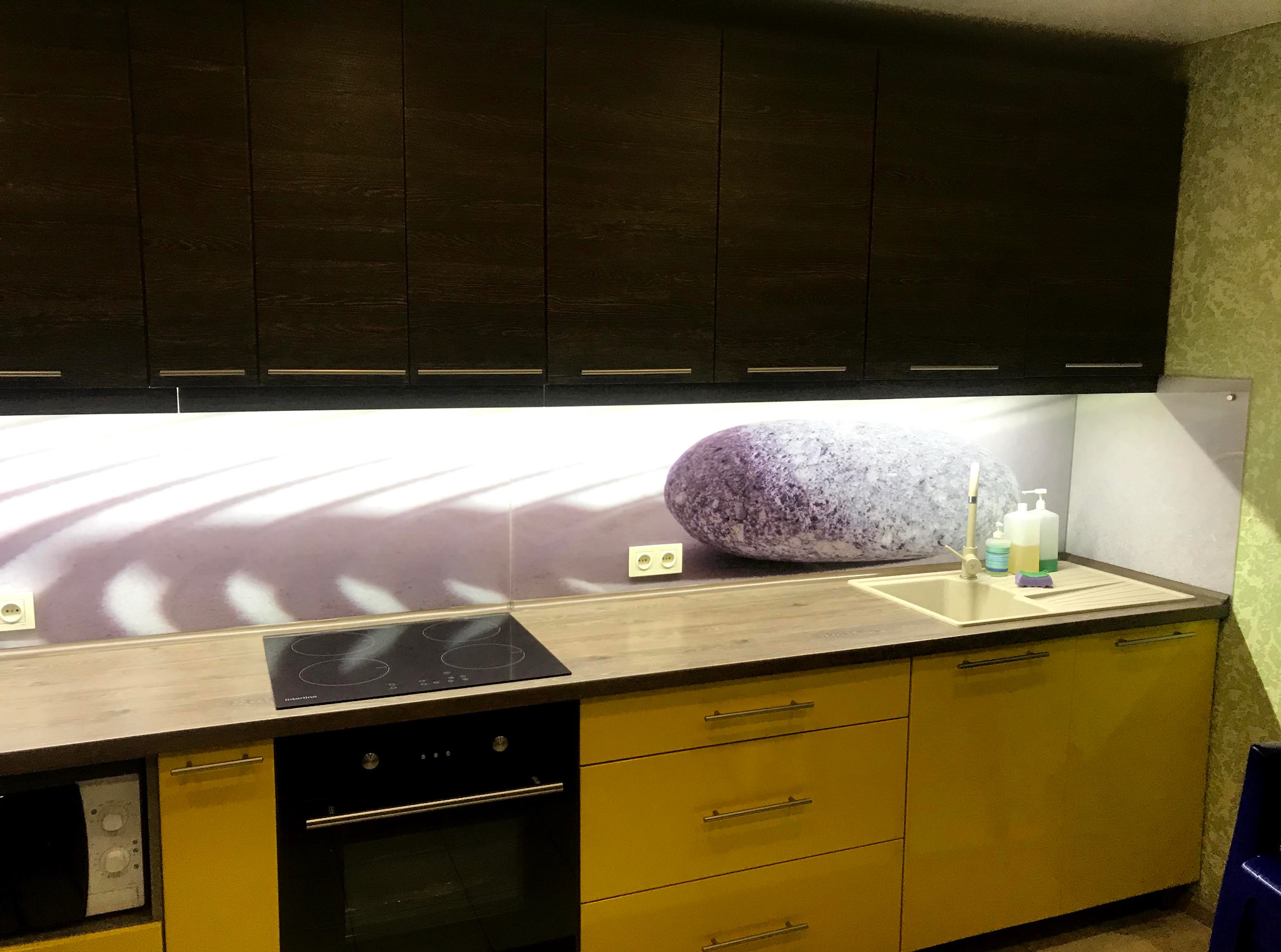 Żółto-czarna kuchnia z blatem imitującym drewno. Na ścianie grafika z motywem kamienia. Widoczna płyta indukcyjna i piekarnik. Górne szafki ciemne, drewnopodobne.