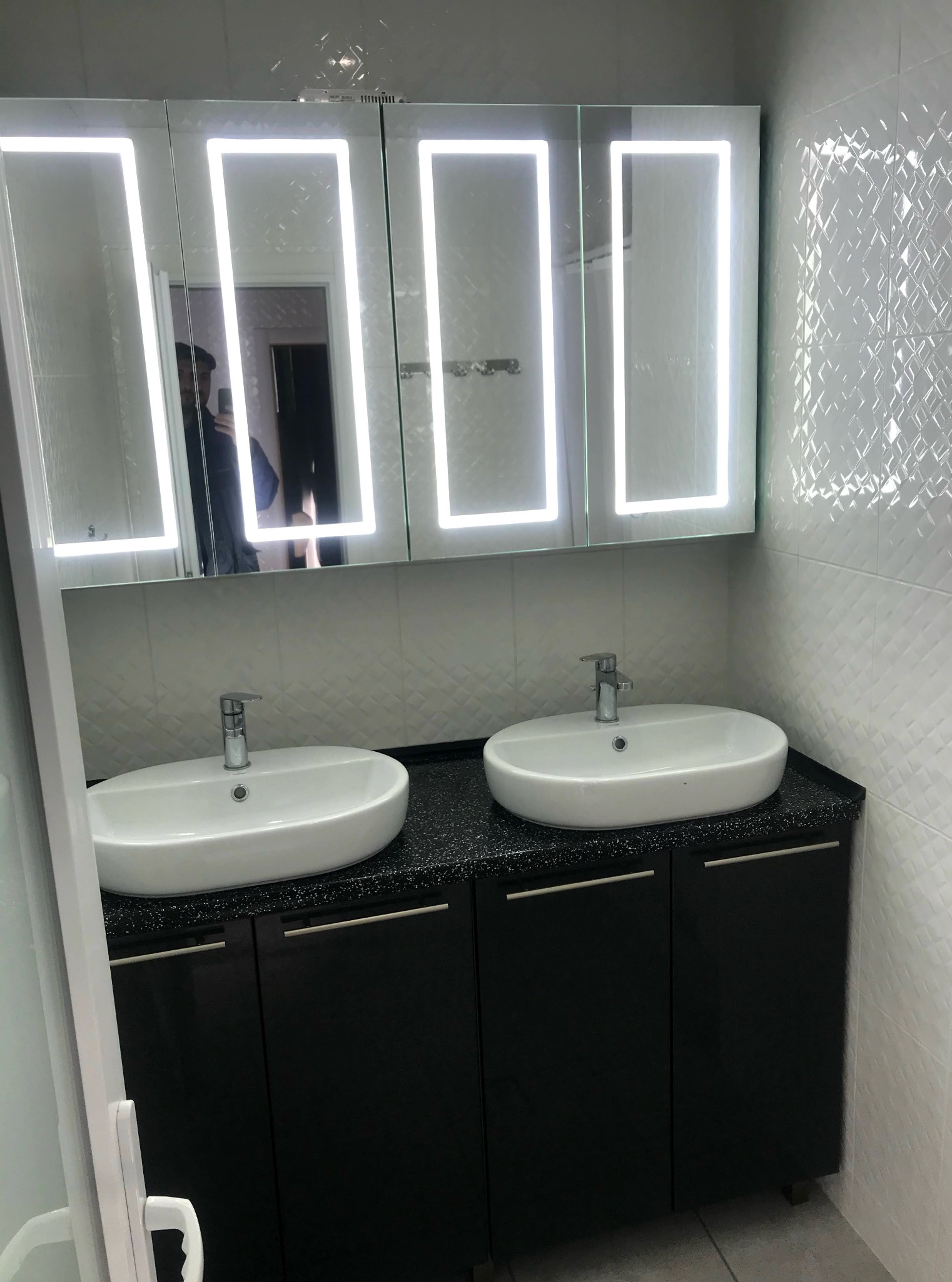 Nowoczesna łazienka z szafką pod umywalkę w kolorze czarnym, umywalkami nablatowymi i lustrem z podświetleniem LED. Białe płytki na ścianie.