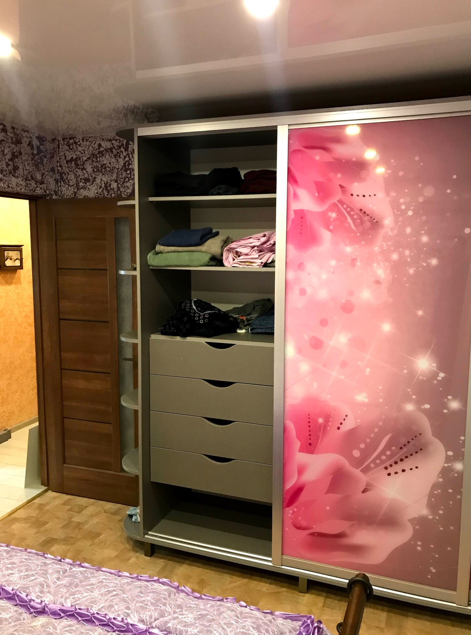 Szafa wnękowa z przesuwnymi drzwiami, zdobiona motywem kwiatowym w odcieniach różu, z widocznymi półkami i szufladami, częściowo wypełniona ubraniami.