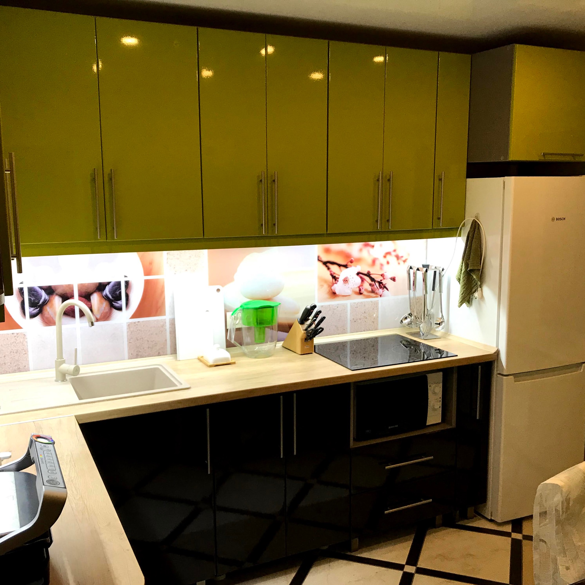 Nowoczesna kuchnia z oliwkowymi szafkami górnymi i czarnymi dolnymi, drewniany blat, wzorzysty panel nad blatem, sprzęt AGD. Całość oświetlona.