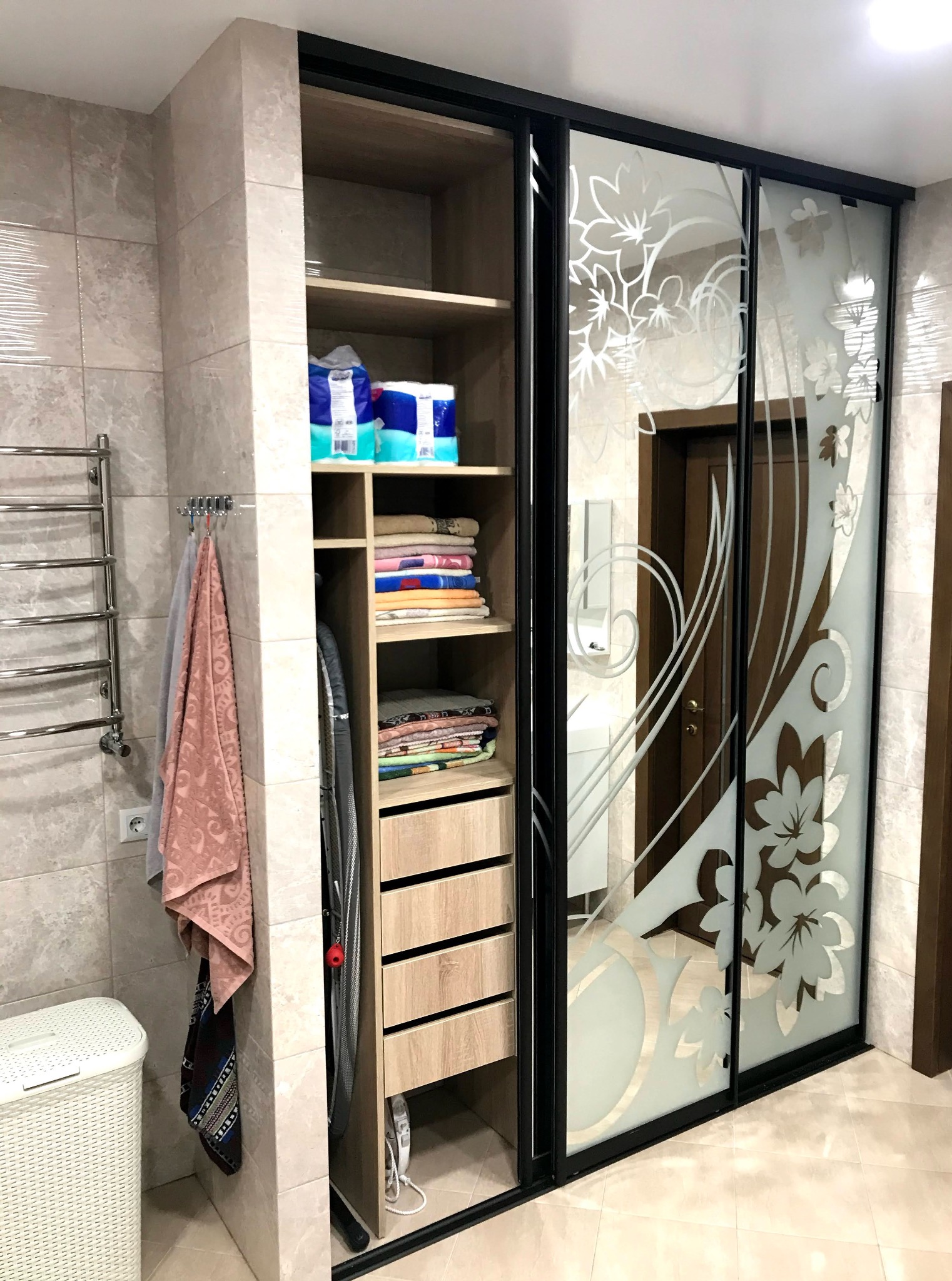 Szafa wnękowa z przesuwnymi drzwiami zdobionymi motywem kwiatowym, w łazience z ręcznikami i grzejnikiem drabinkowym, widok na wnętrze szafy z półkami i szufladami.