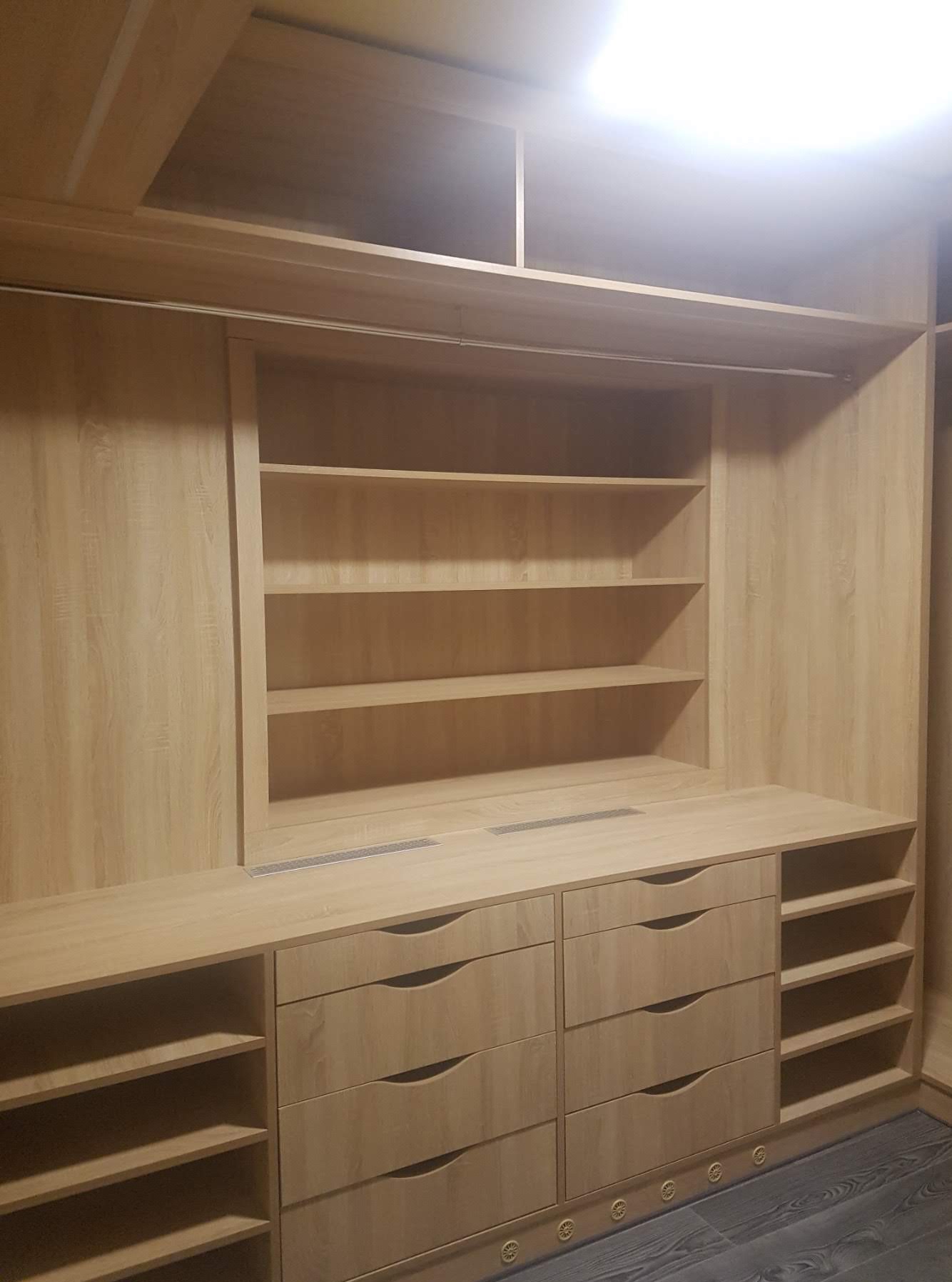 Jasna, drewniana szafa wnękowa z szufladami i półkami, oświetlona od góry. Widoczna faktura drewna i metalowe detale wentylacyjne na blacie.