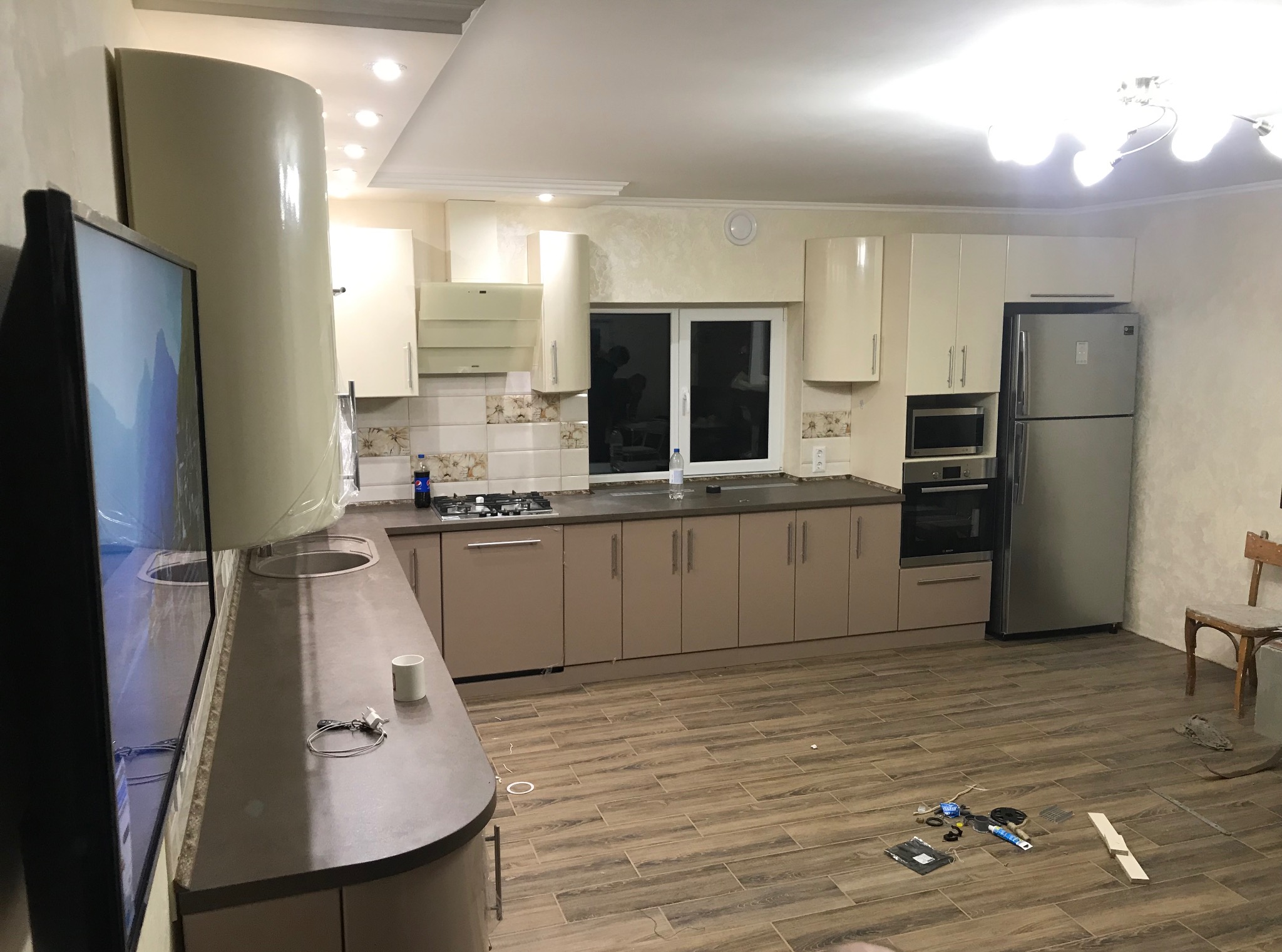 Jasna kuchnia z beżowymi szafkami, brązowym blatem i drewnopodobną podłogą. Na podłodze leżą narzędzia i akcesoria montażowe. Widoczne okno i krzesło.