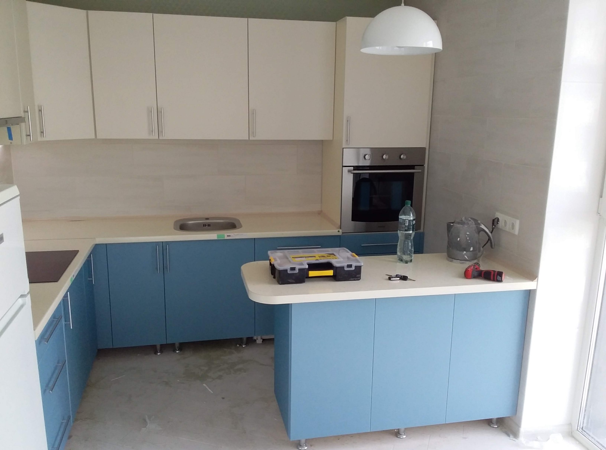 Jasna kuchnia z niebieskimi dolnymi szafkami i białymi górnymi, z narzędziami na blacie. Widoczny piekarnik i biała lampa sufitowa.