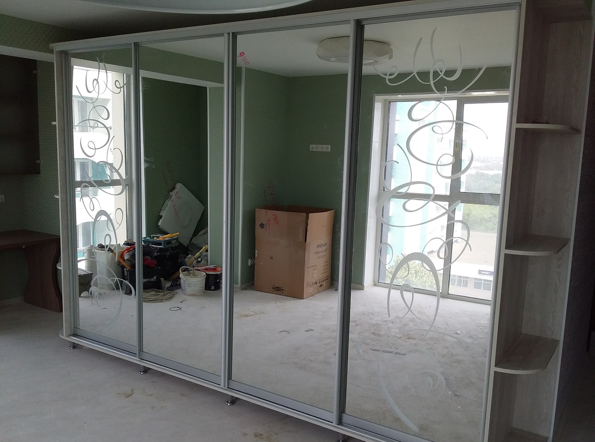 Szafa przesuwna z lustrami zdobionymi wzorem, widok na wnętrze pokoju z kartonem i narzędziami, jasne wykończenie, metalowe ramy drzwi.