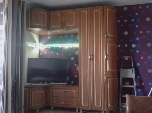 Funkcjonalna szafa wnękowa z segmentem RTV w kolorze brązu, z dekoracyjnymi frezami, oświetleniem LED i telewizorem na tle ściany w fioletowe wzory.