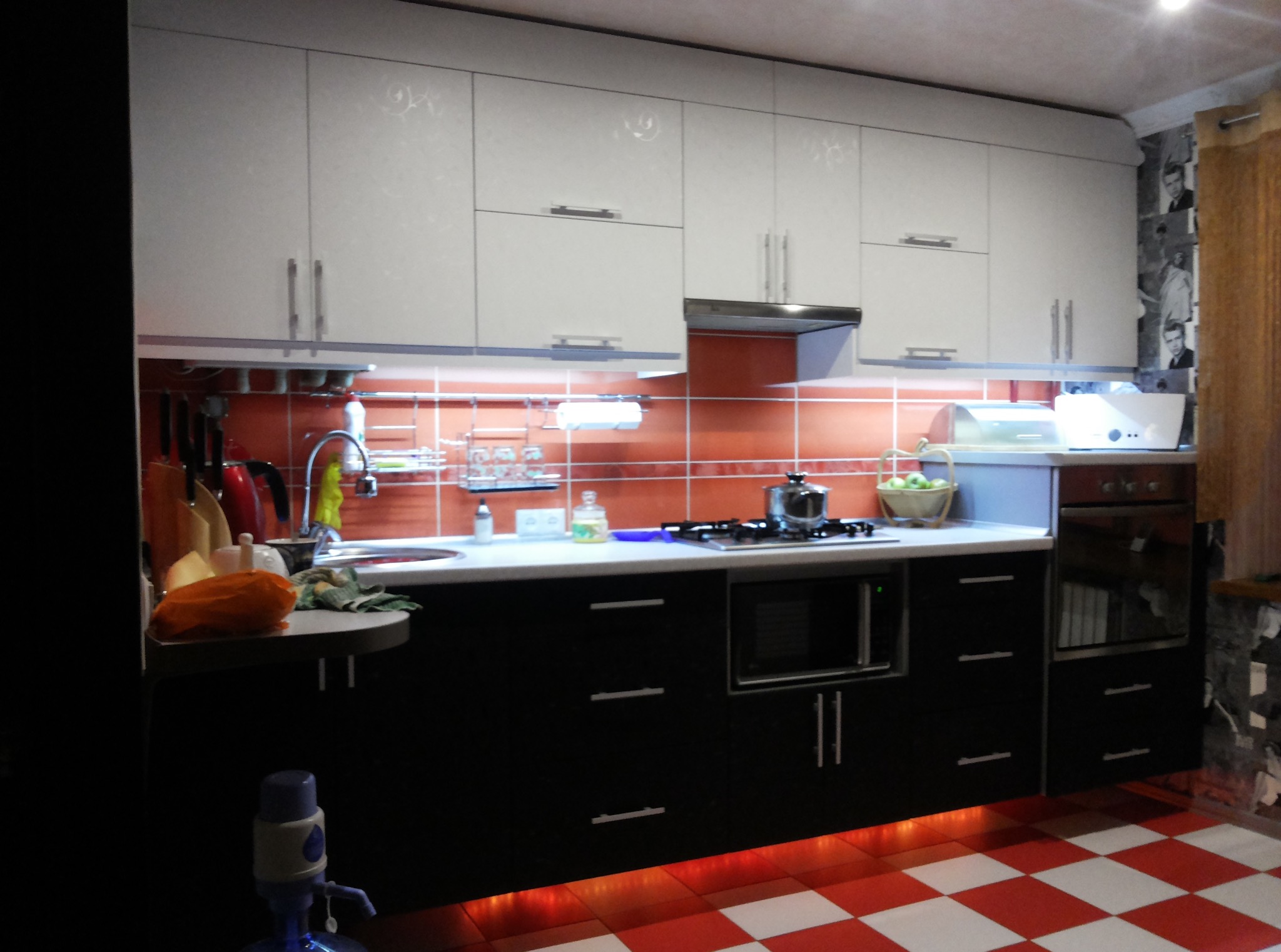 Nowoczesna kuchnia z białymi szafkami górnymi i czarnymi dolnymi, oświetleniem LED pod szafkami i czerwoną mozaiką na ścianie oraz podłodze w szachownicę.