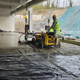 Wylewka posadzki przemysłowej w Boruja Kościelna wykonywana maszynowo z użyciem listwy wibracyjnej na folii. Widoczny pracownik w żółtej kamizelce.