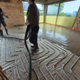 Wylewka anhydrytowa w trakcie realizacji: pracownicy rozprowadzają płynny jastrych na ogrzewaniu podłogowym w surowym wnętrzu budynku z ceglanymi ścianami.