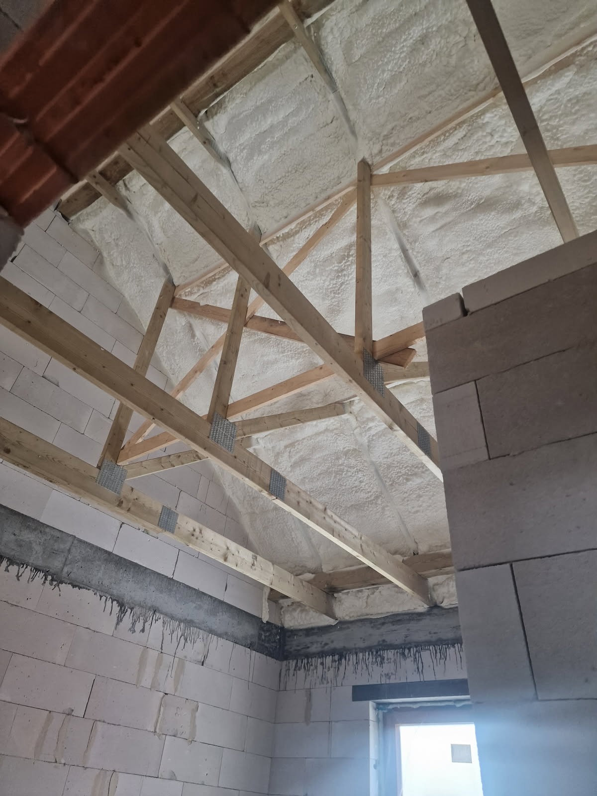 Poddasze z drewnianą konstrukcją pokryte białą pianą PUR, widok z dołu, fragment ściany z betonu komórkowego po prawej. Ocieplenie natryskowe.
