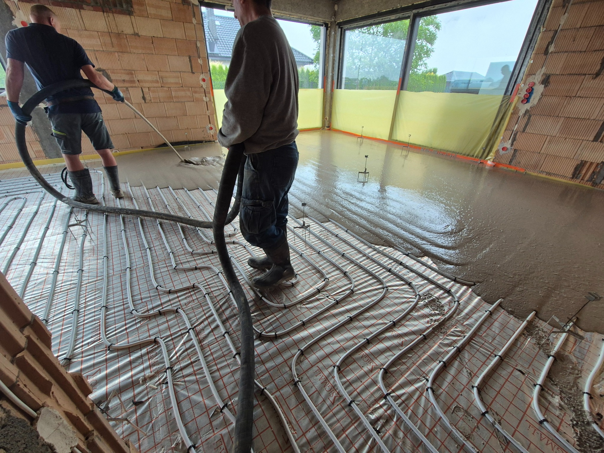 Wylewka anhydrytowa w trakcie realizacji: pracownicy rozprowadzają płynny jastrych na ogrzewaniu podłogowym w surowym wnętrzu budynku z ceglanymi ścianami.