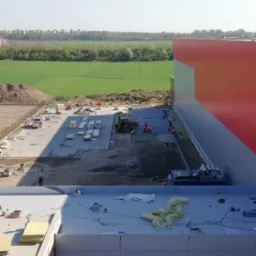 Budowa hali przemysłowej: widok z góry na plac budowy z materiałami, maszynami i pracownikami. W tle zielone pola i zabudowa miejska.