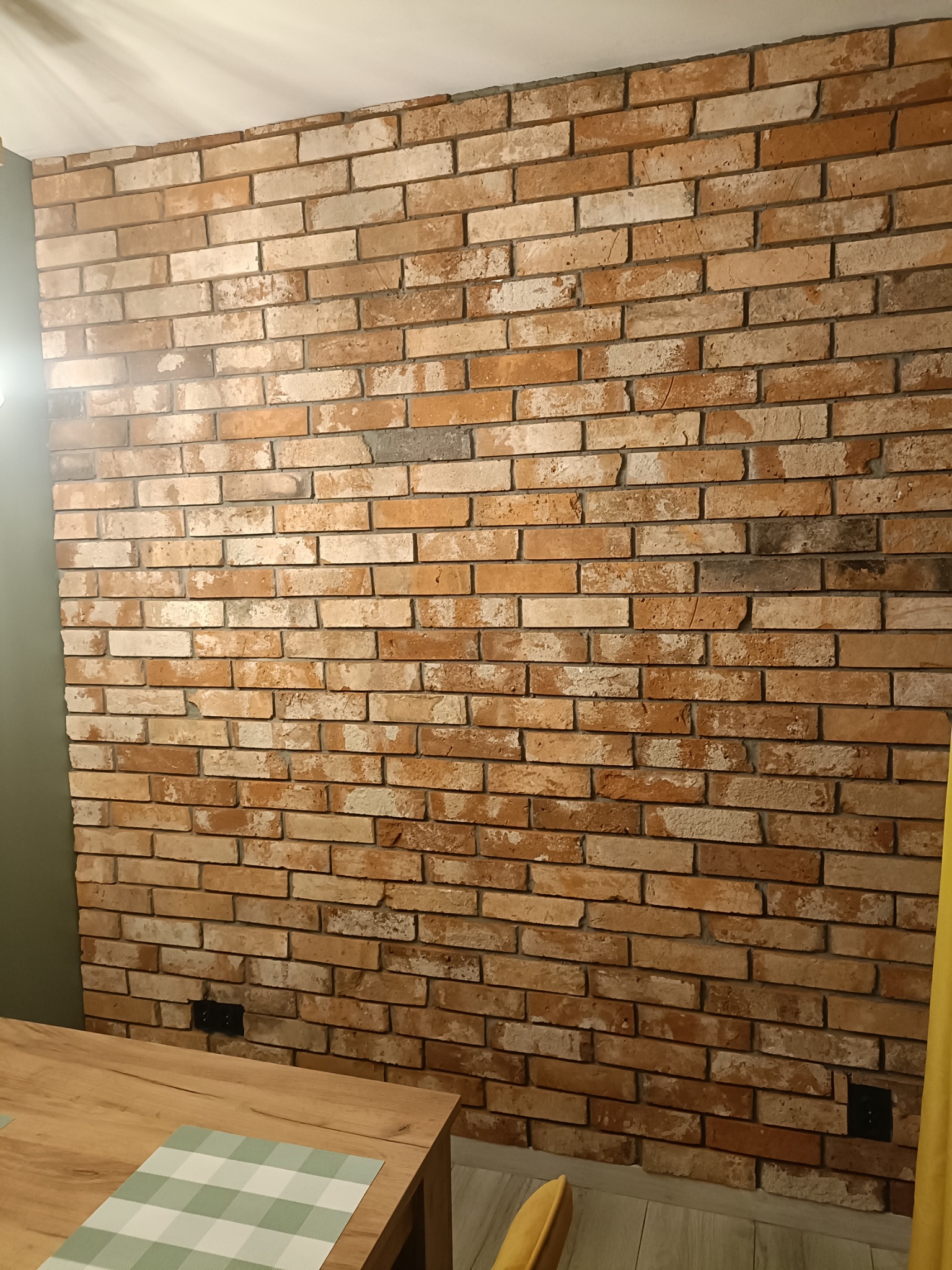 Ściana z cegły dekoracyjnej w salonie, oświetlona z góry. Widoczny fragment stołu z nakryciem. Cegła w odcieniach brązu, beżu i szarości. Gniazdka elektryczne w ścianie.