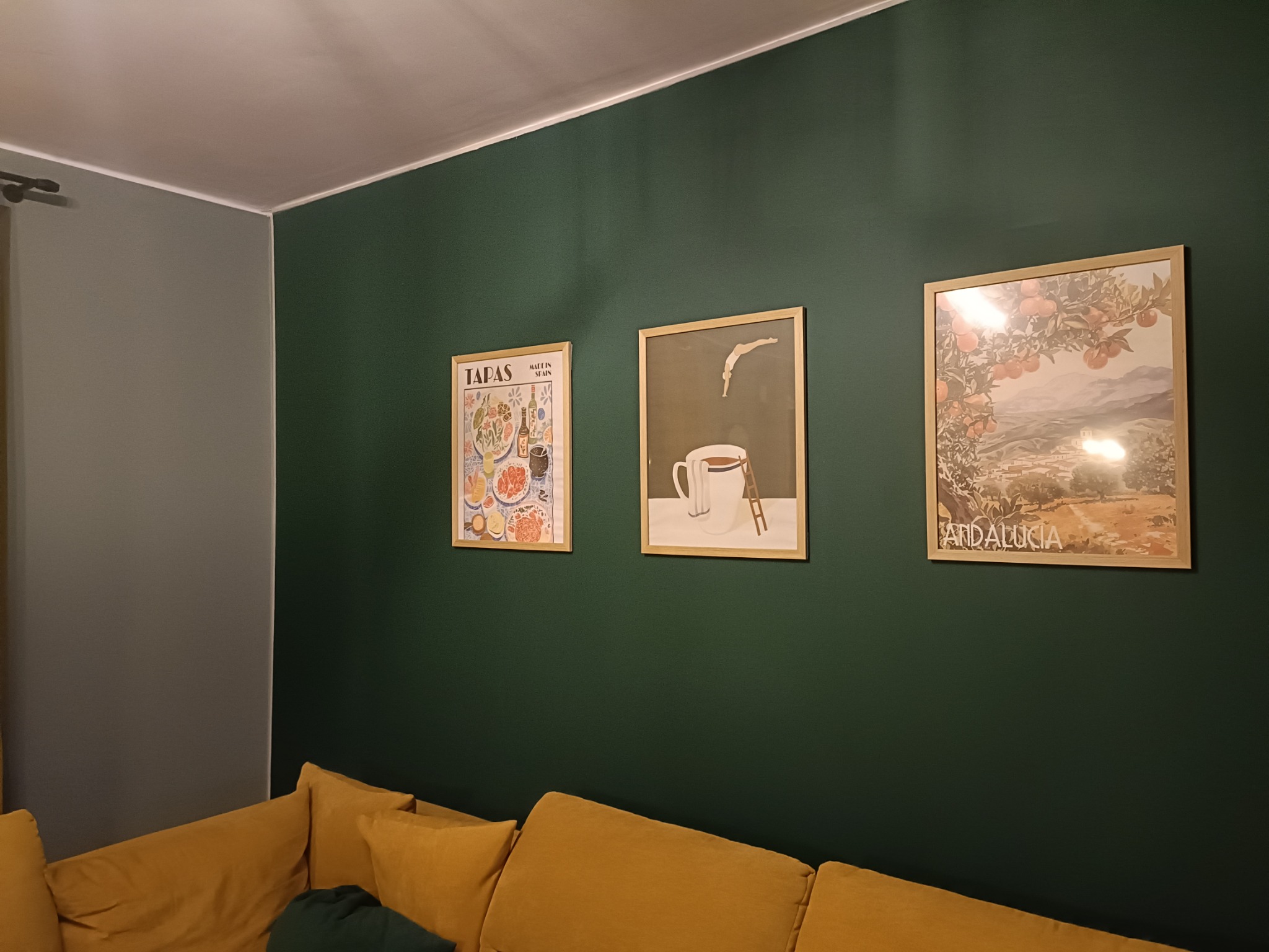Ciemnozielona ściana z trzema oprawionymi plakatami w jasnych ramkach nad żółtą sofą. Szara ściana z lewej strony kadru. Minimalistyczny wystrój wnętrza.