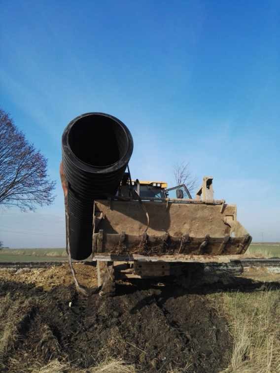 Koparka transportuje grubą rurę drenarską na tle błękitnego nieba. Widoczny krajobraz polny i fragment drzewa.
