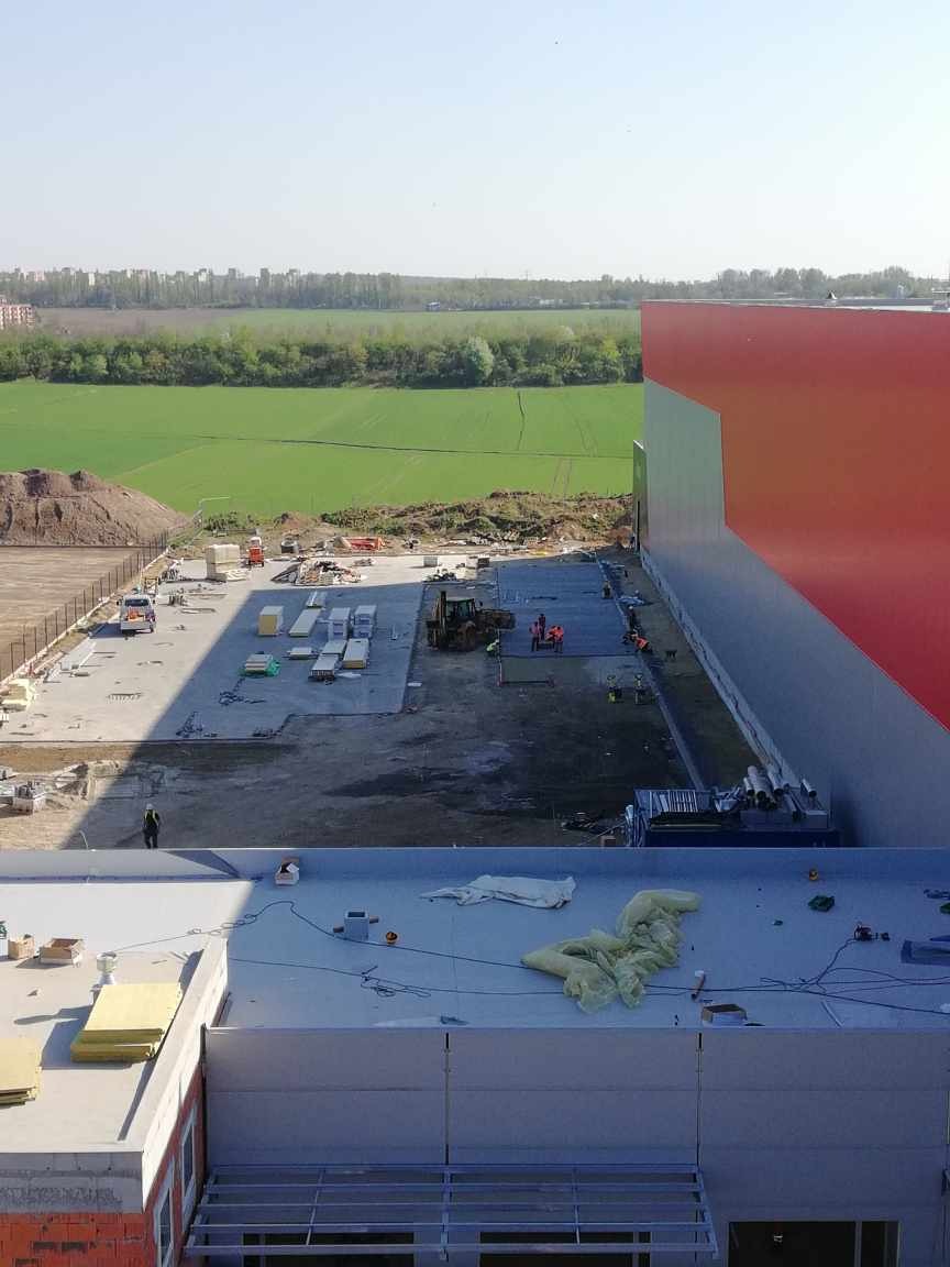 Budowa hali przemysłowej: widok z góry na plac budowy z materiałami, maszynami i pracownikami. W tle zielone pola i zabudowa miejska.