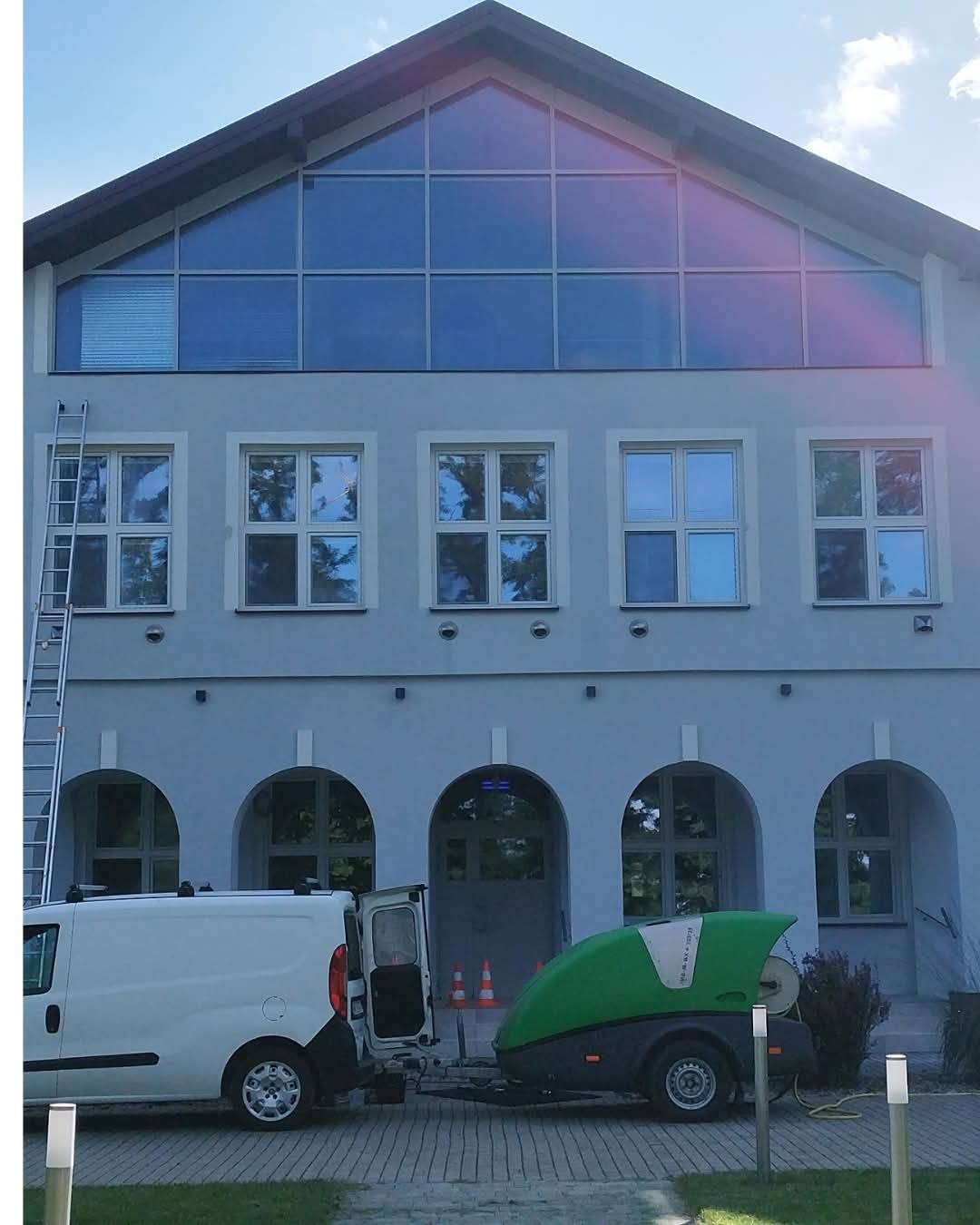 Budynek z szarymi ścianami i dużymi oknami, drabina oparta o ścianę, biały van z otwartymi drzwiami i przyczepa z myjką ciśnieniową. Usługa czyszczenia elewacji.