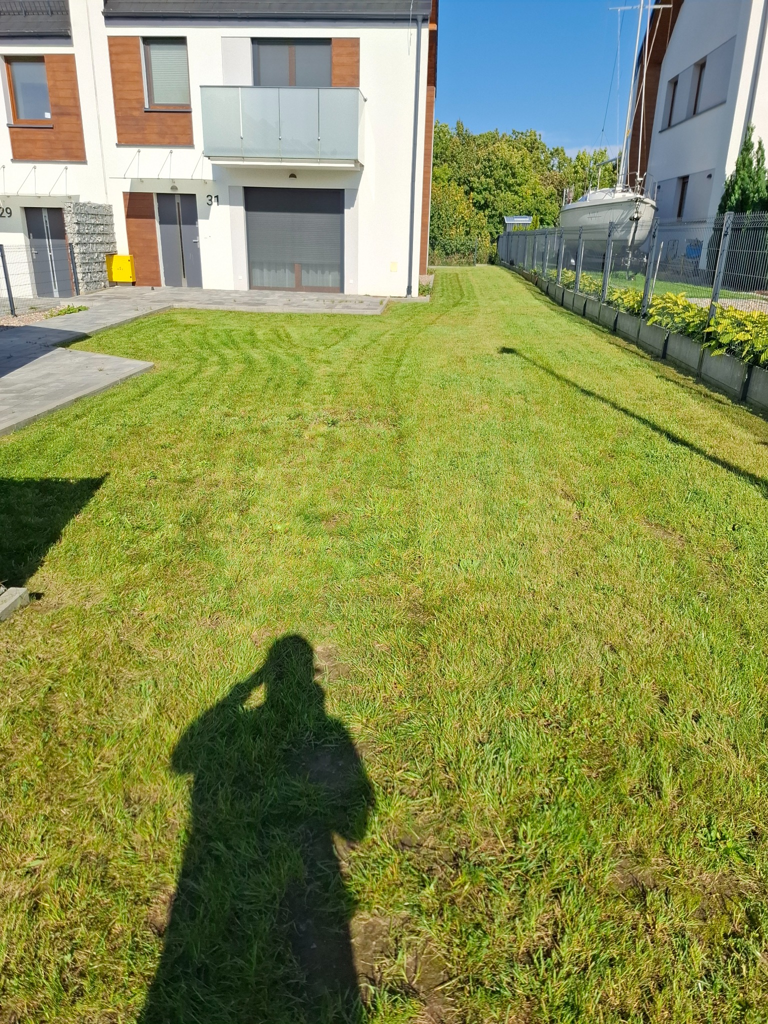Świeżo skoszony trawnik przed szeregową zabudową, z widocznym cieniem osoby fotografującej. W tle zaparkowany jacht za ogrodzeniem.