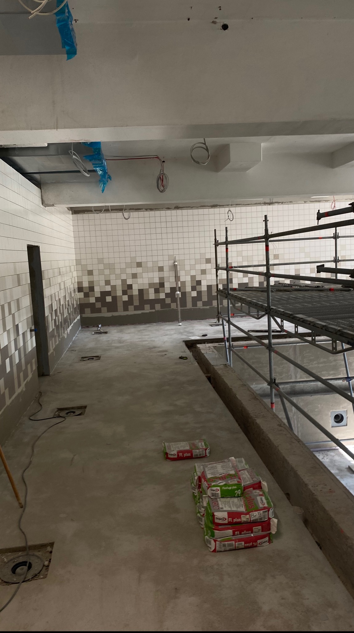 Wnętrze w trakcie prac: ułożone płytki na ścianach w mozaikę, worki z zaprawą na podłodze, rusztowania. Widoczne instalacje pod sufitem.
