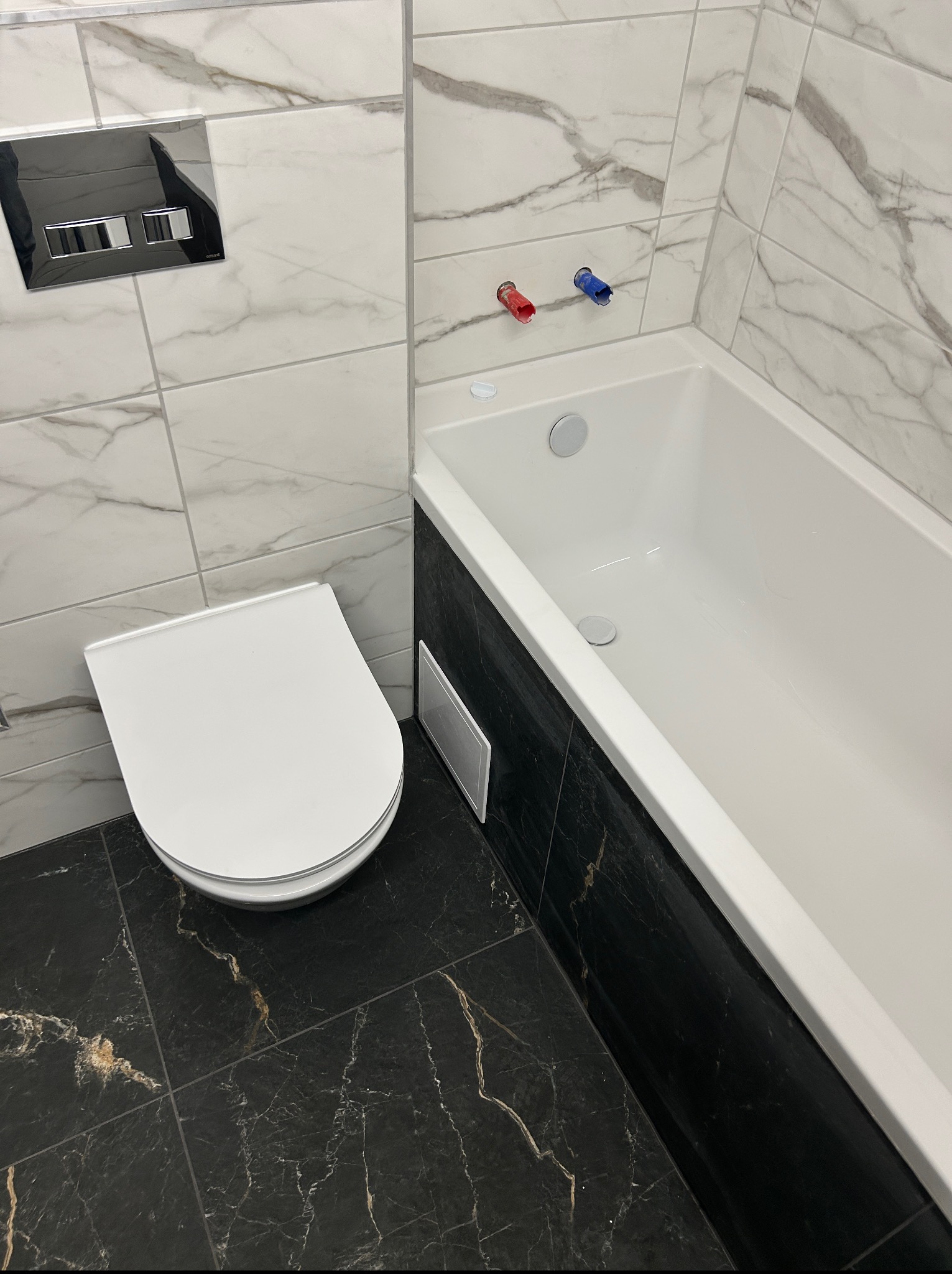 Wykończona łazienka: biała wanna, podwieszana toaleta i czarne płytki podłogowe z marmurkowym wzorem. Białe płytki ścienne z szarymi żyłami imitują marmur.