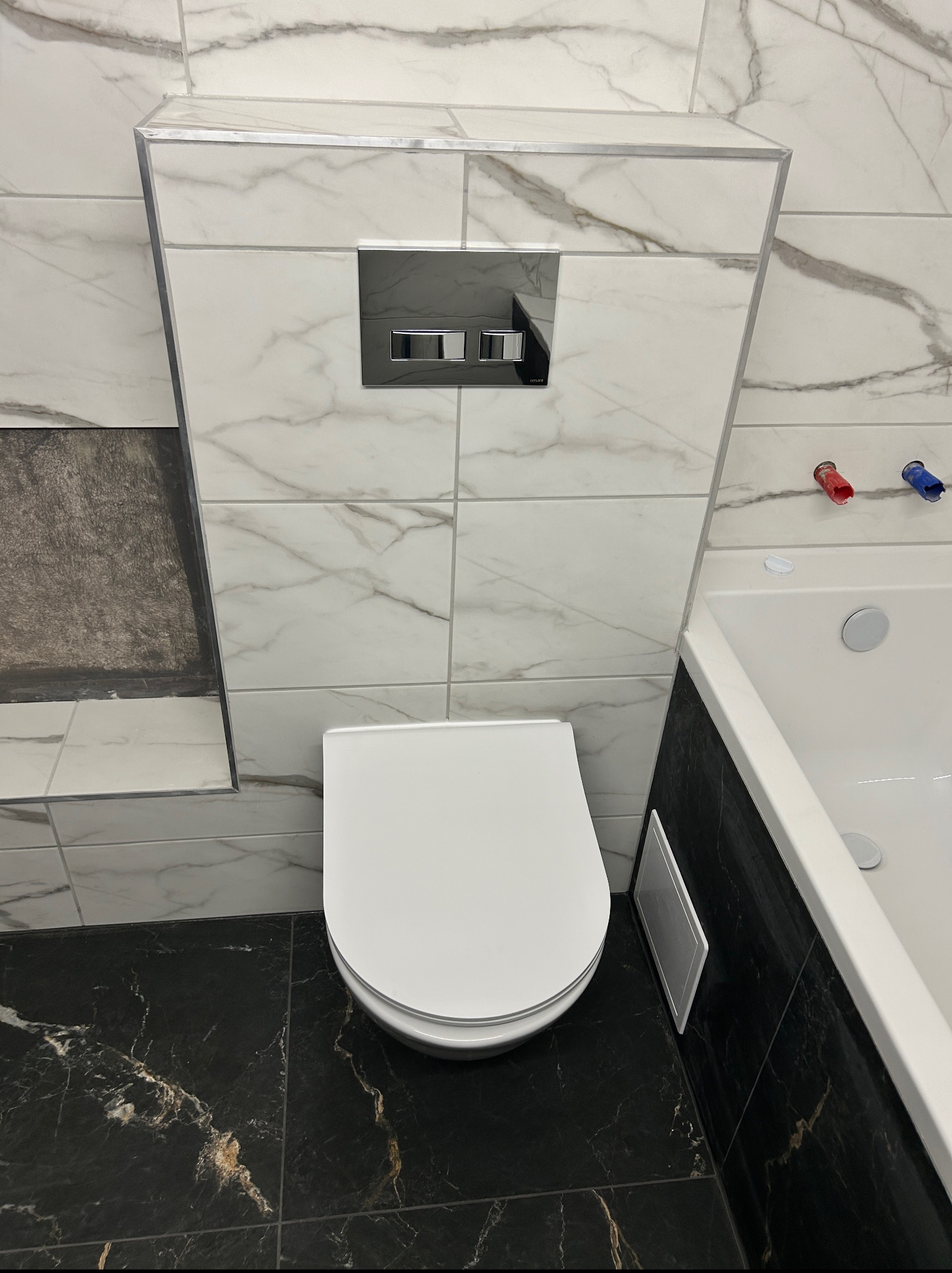 Nowoczesna toaleta podwieszana z chromowanym przyciskiem spłukującym, obudowana płytkami imitującymi marmur. Czarne płytki podłogowe z wyraźnym, złotym żyłowaniem.