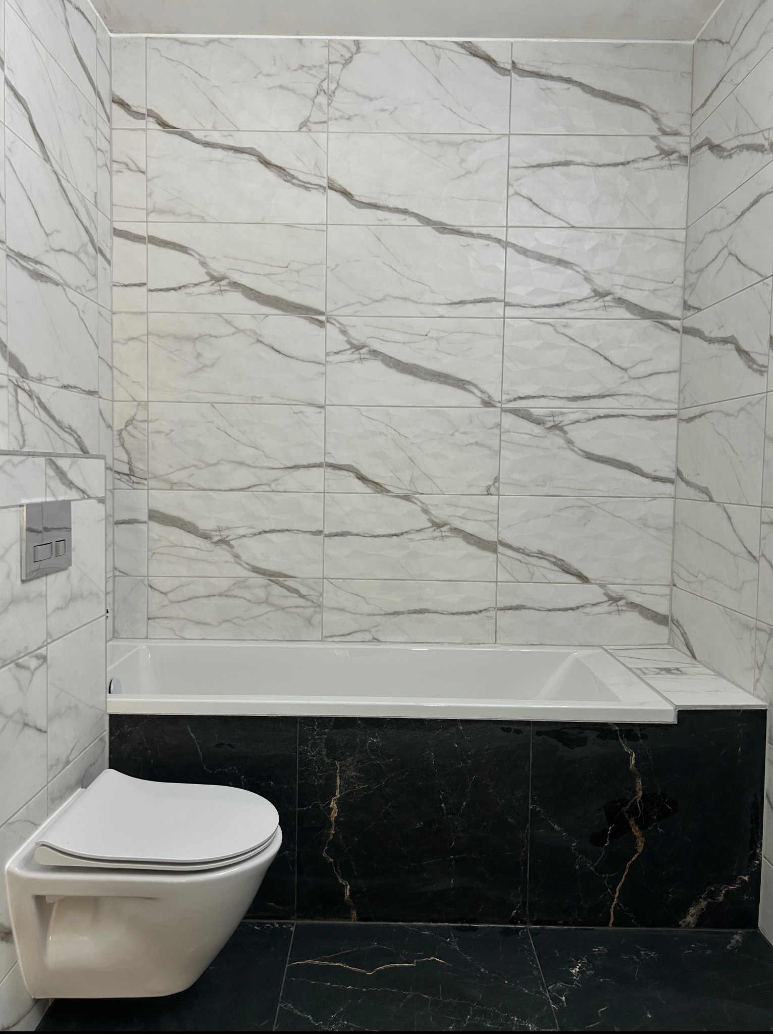 Wykończona łazienka: białe płytki marmurowe na ścianach, czarne płytki marmurowe na podłodze i obudowie wanny, podwieszana toaleta z chromowanym przyciskiem.