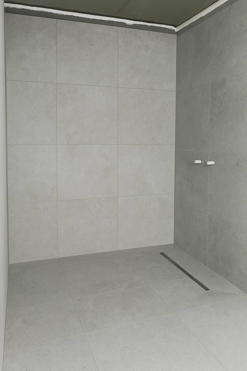 Wykończona, minimalistyczna kabina prysznicowa z szarymi, kwadratowymi płytkami i odpływem liniowym. Widoczne dwa uchwyty na ścianie.