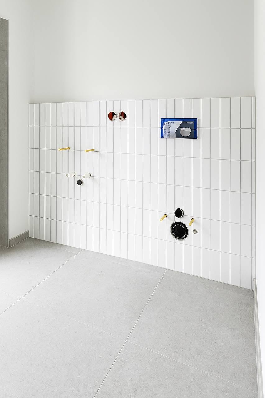 Białe, pionowe płytki w łazience z widocznymi instalacjami i otworami na armaturę. Szare płytki podłogowe i minimalistyczny styl.