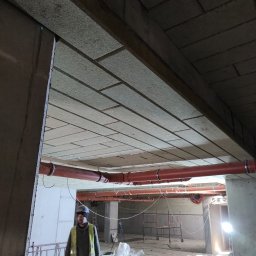 Vlas-bud Volodymyr Vlasenko - Ocieplony strop garażu podziemnego z widocznymi rurami i pracownikiem w kamizelce odblaskowej, ujęcie z dołu, w tle rusztowania i worki z materiałami.