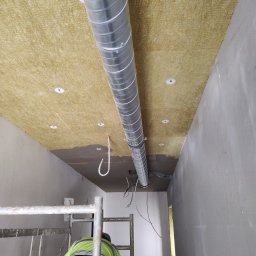 Vlas-bud Volodymyr Vlasenko - Ocieplenie sufitu wełną mineralną w korytarzu, widoczny fragment instalacji wentylacyjnej i pracownik na rusztowaniu. Etap klejenia wełny i montażu kołków.