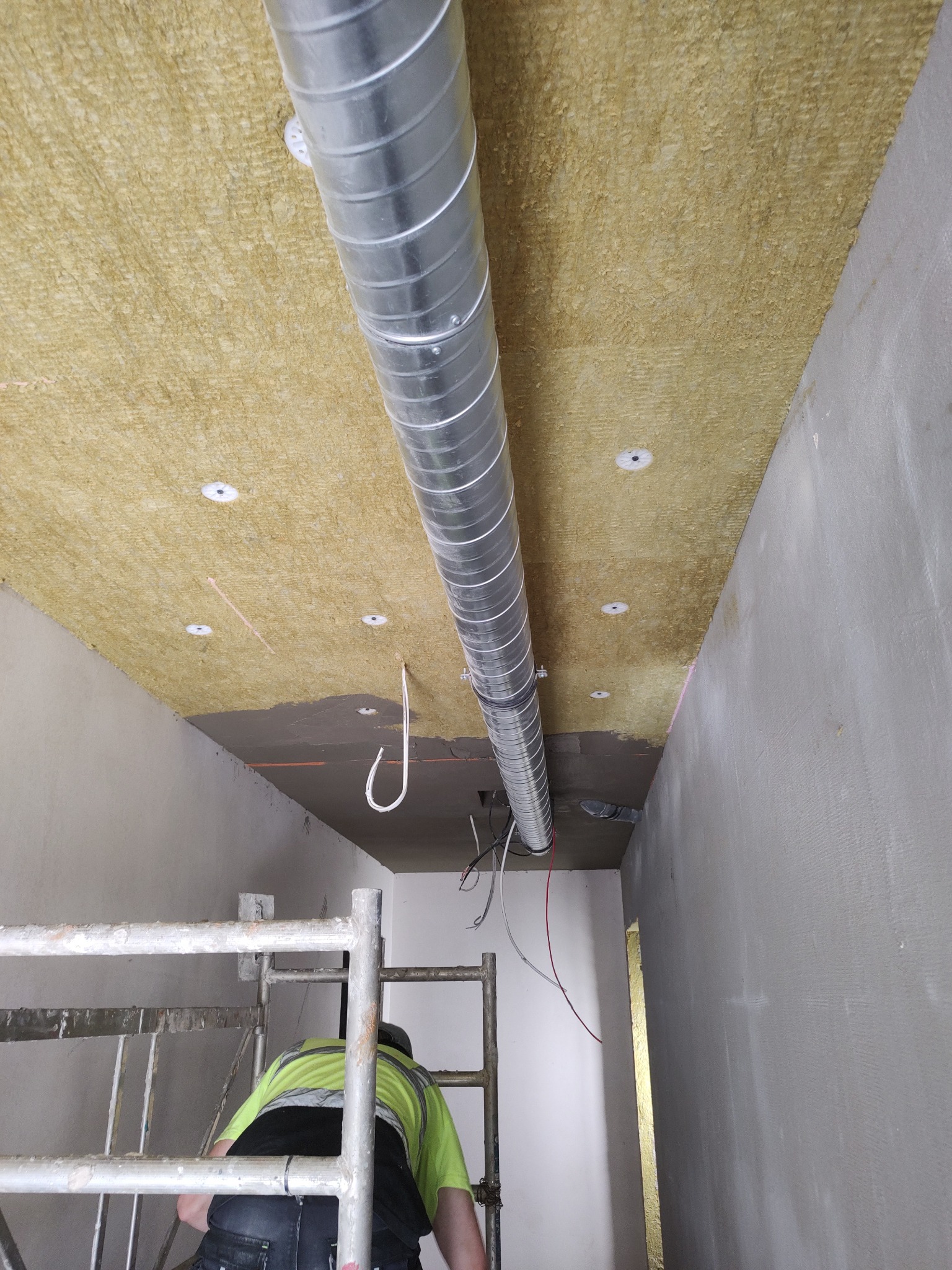 Ocieplenie sufitu wełną mineralną w korytarzu, widoczny fragment instalacji wentylacyjnej i pracownik na rusztowaniu. Etap klejenia wełny i montażu kołków.