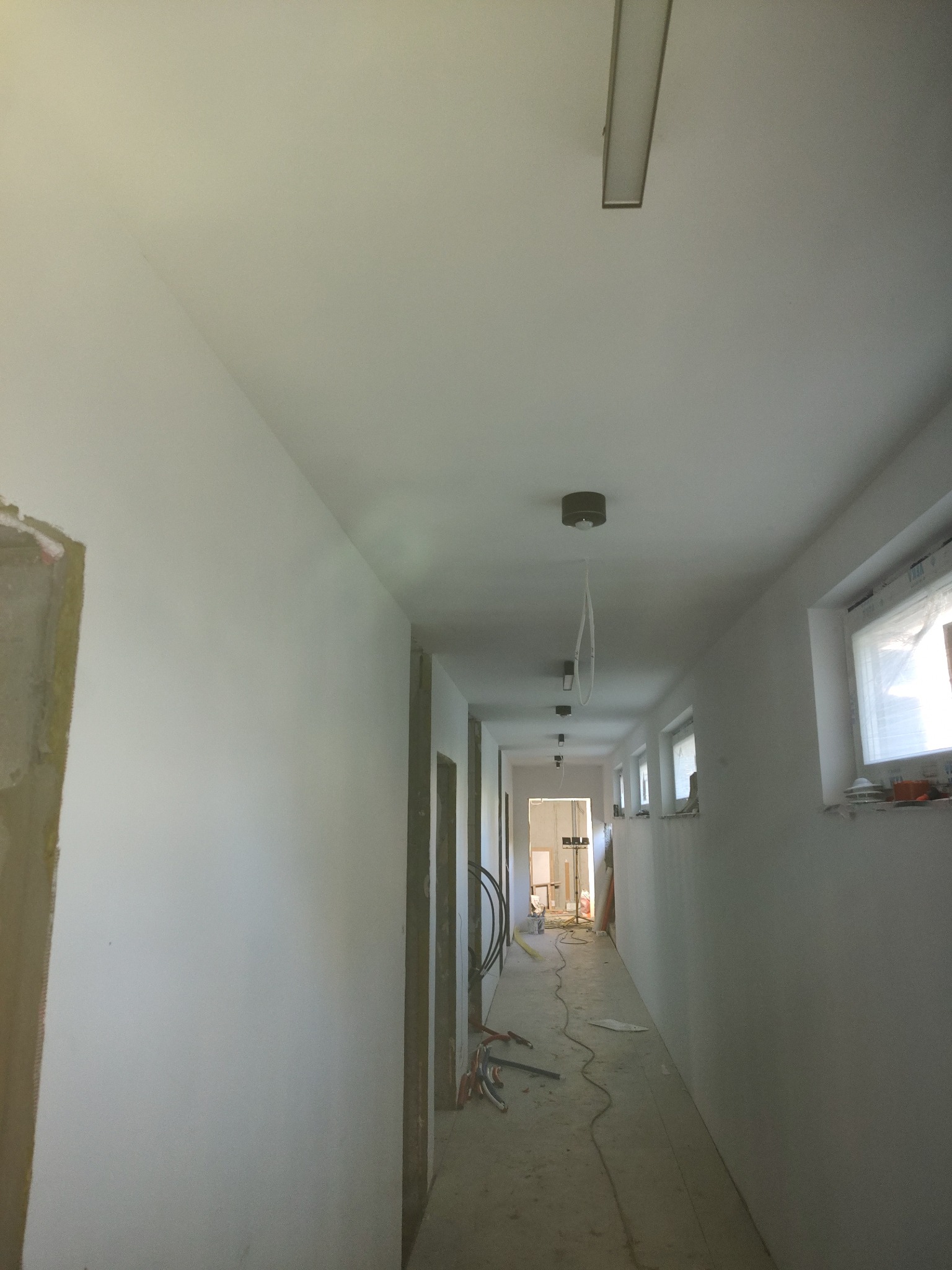 Biały korytarz w bloku po gładziowaniu i malowaniu podkładowym; widoczne okna, lampy, narzędzia i surowa ościeżnica.